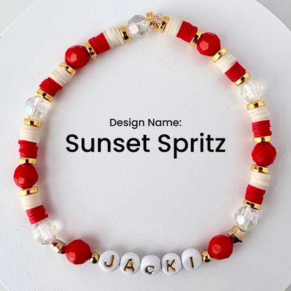 A beaded bracelet with the text "Sunset Spritz." The bracelet features red, white, gold, and clear beads. The name "JACKI" is spelled out in white letter beads. The bracelet is designed with a mix of round and disc-shaped beads.