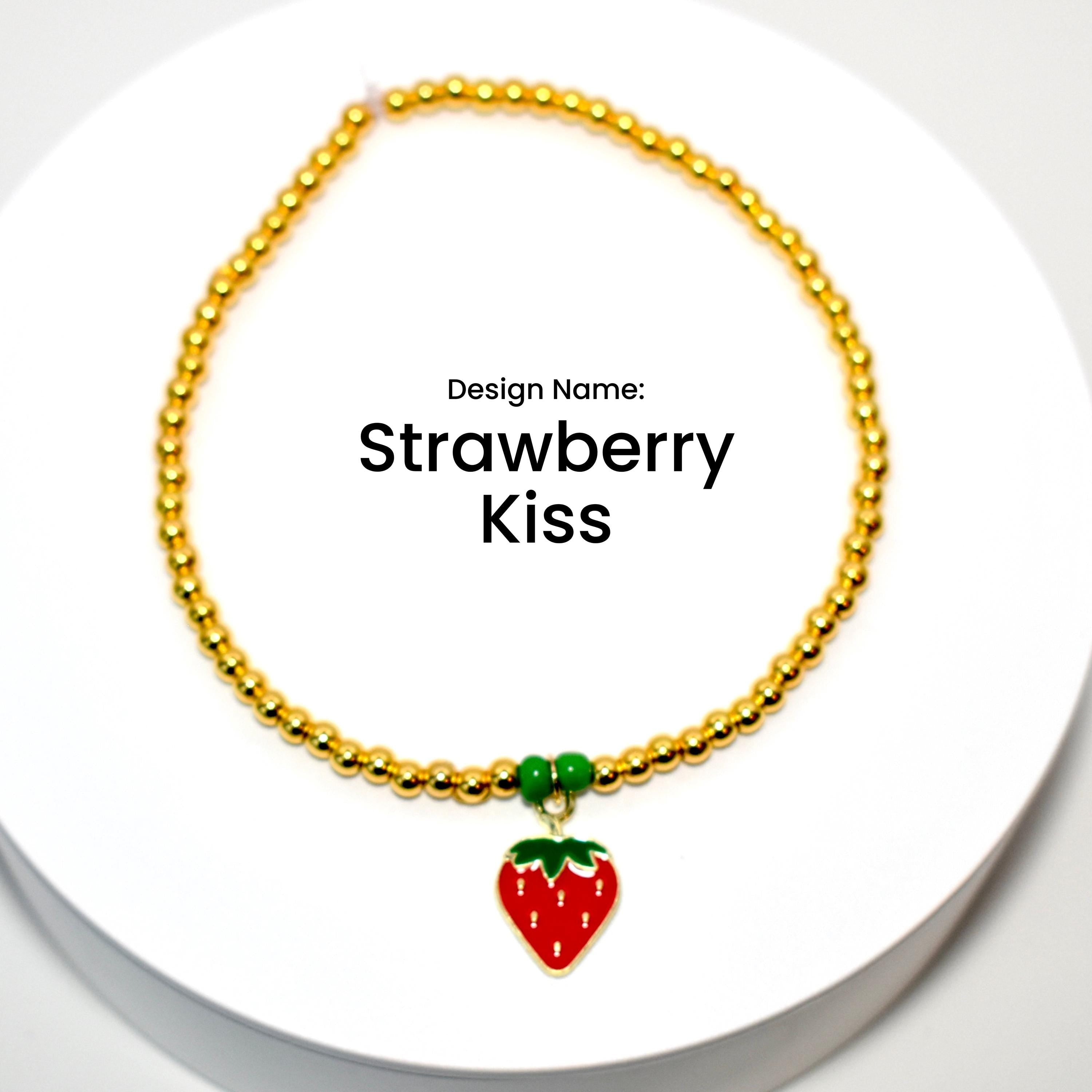 A gold beaded bracelet with a red and green strawberry charm. The bracelet has a design name of 'Strawberry Kiss'.