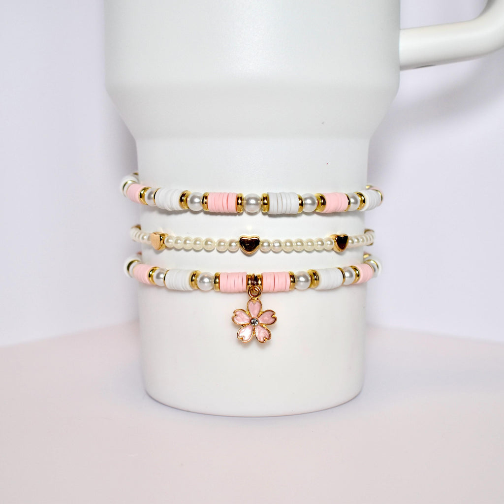 Three layered bracelets with pink and white beads and gold accents. The bottom bracelet has a pink flower charm.