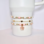 Three layered bracelets with pink and white beads and gold accents. The bottom bracelet has a pink flower charm.