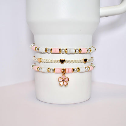 Three layered bracelets with pink and white beads and gold accents. The bottom bracelet has a pink flower charm.