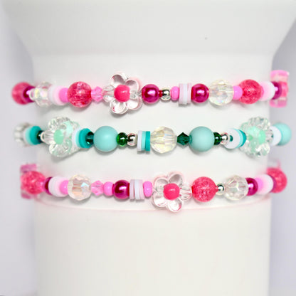Three pink, turquoise, and white beaded bracelets with flower charms. The bracelets are stacked on top of each other.