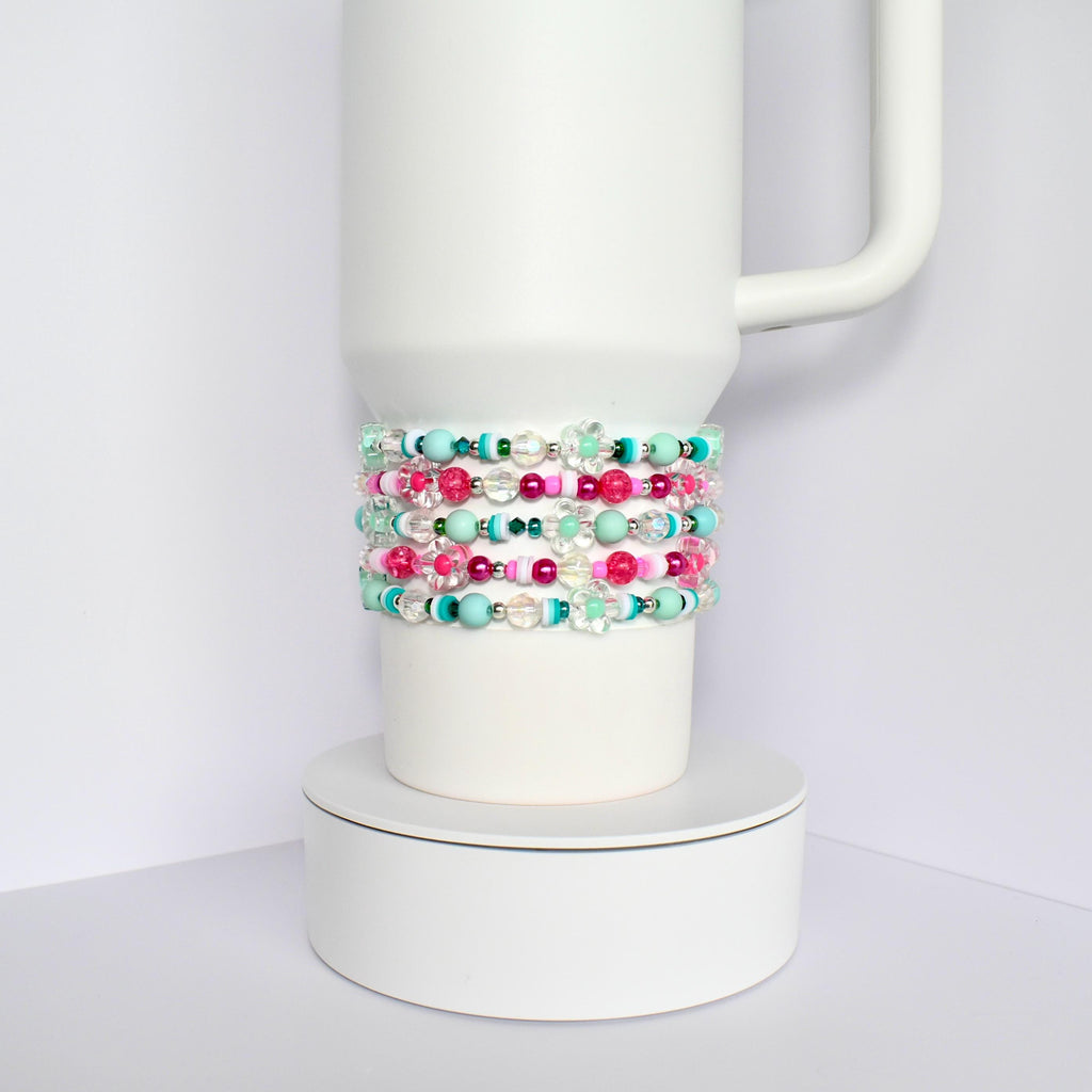 A white tumbler with a stack of colorful beaded bracelets. The bracelets are made of pink, green, blue, and clear beads.