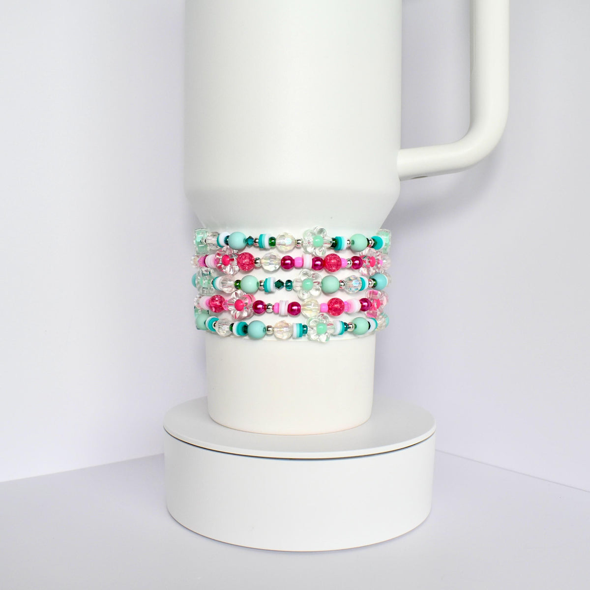 A white tumbler with a stack of colorful beaded bracelets. The bracelets are made of pink, green, blue, and clear beads.