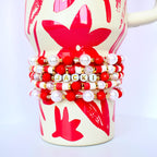 A stack of beaded bracelets in red, white, gold, and pearl colors, wrapped around a cream-colored mug with a red floral design. One bracelet spells out the name "JACKI" in gold letters.