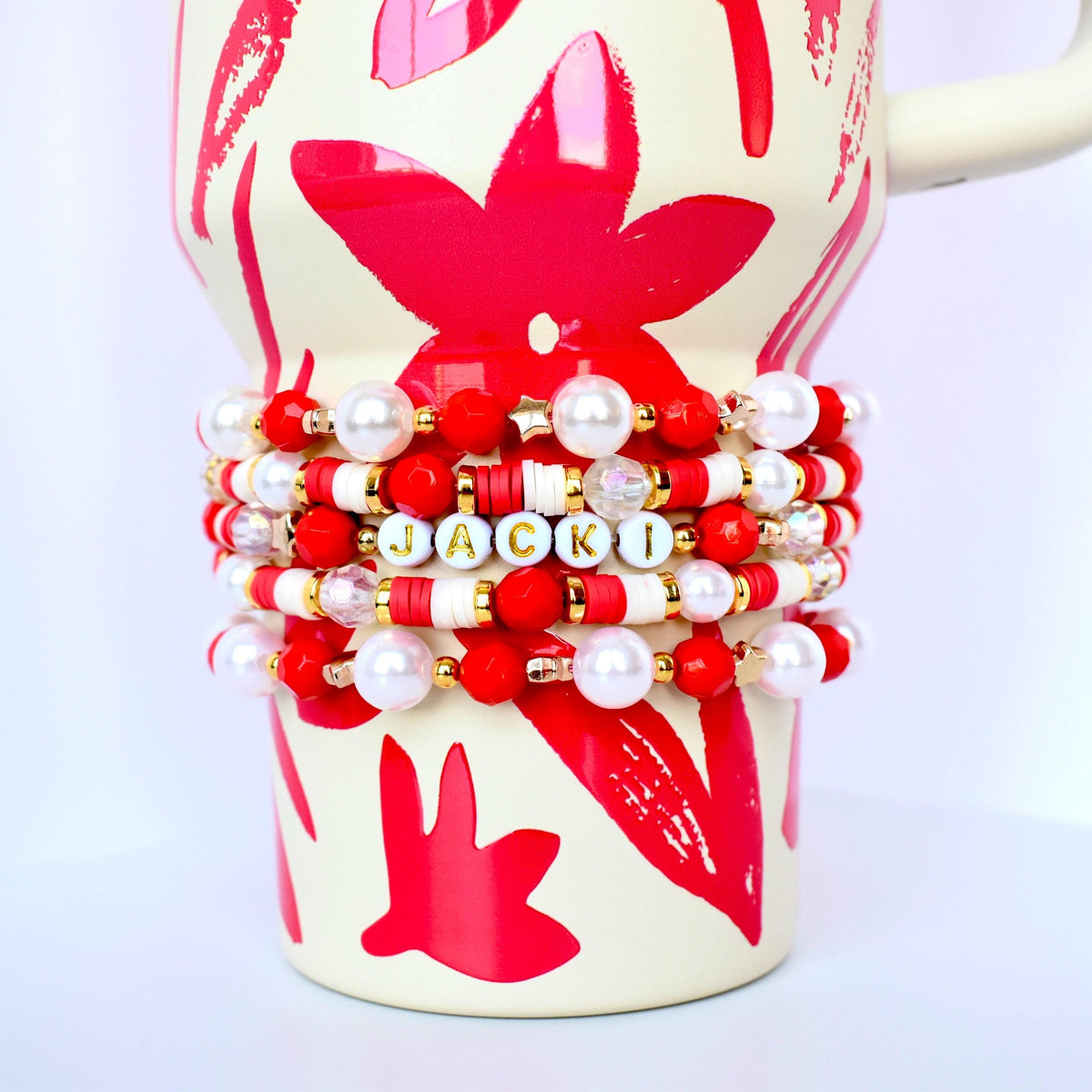 A stack of beaded bracelets in red, white, gold, and pearl colors, wrapped around a cream-colored mug with a red floral design. One bracelet spells out the name "JACKI" in gold letters.