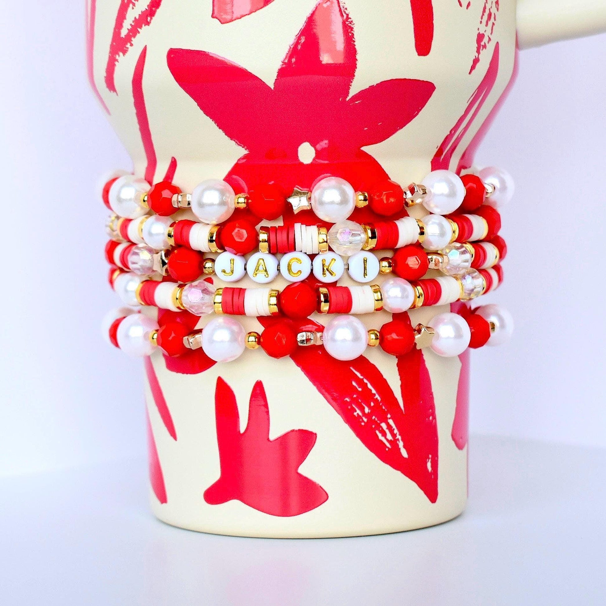 A stack of beaded bracelets in red, white, and gold, with the name "JACKI" spelled out in letter beads. The bracelets are displayed on a cream-colored mug with red floral designs.