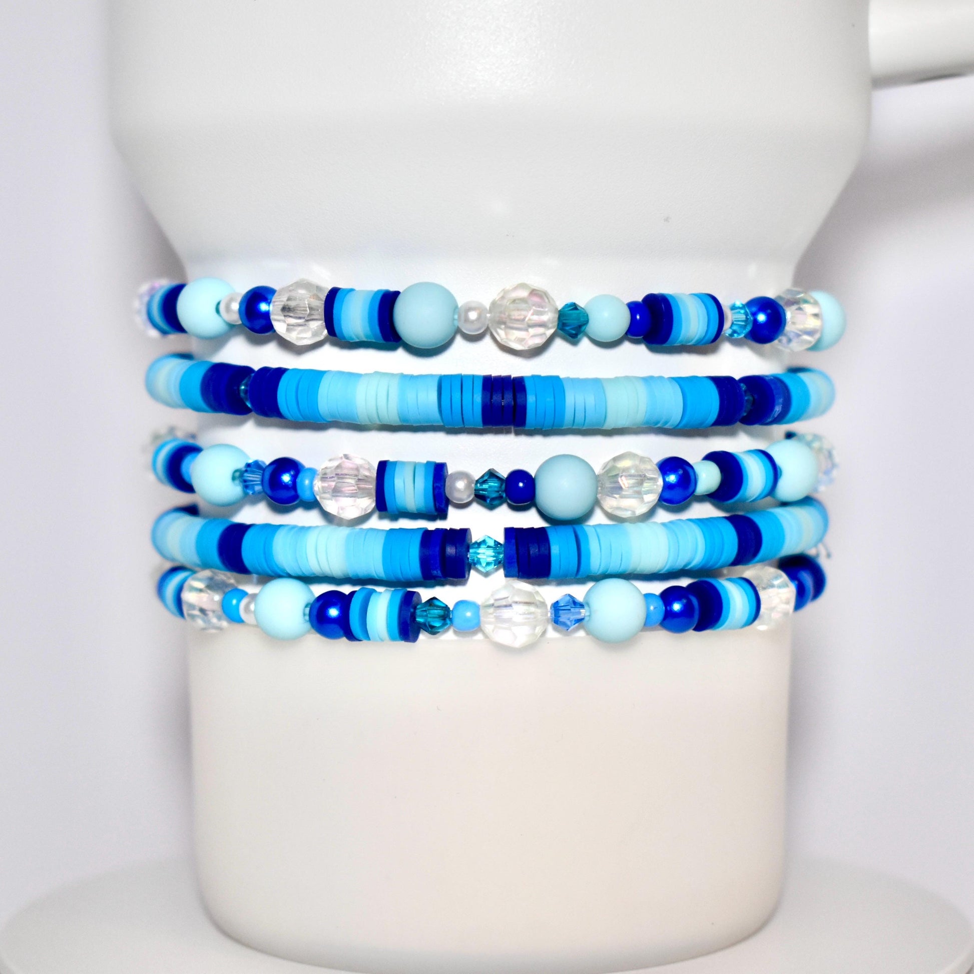 Stack of handmade beaded tumbler bracelets in various shades of blue, including matte, glossy, and translucent beads, wrapped around a white display stand. The Stanley bracelets feature round, disc, and faceted beads in a mix of light blue, dark blue, and clear tones.