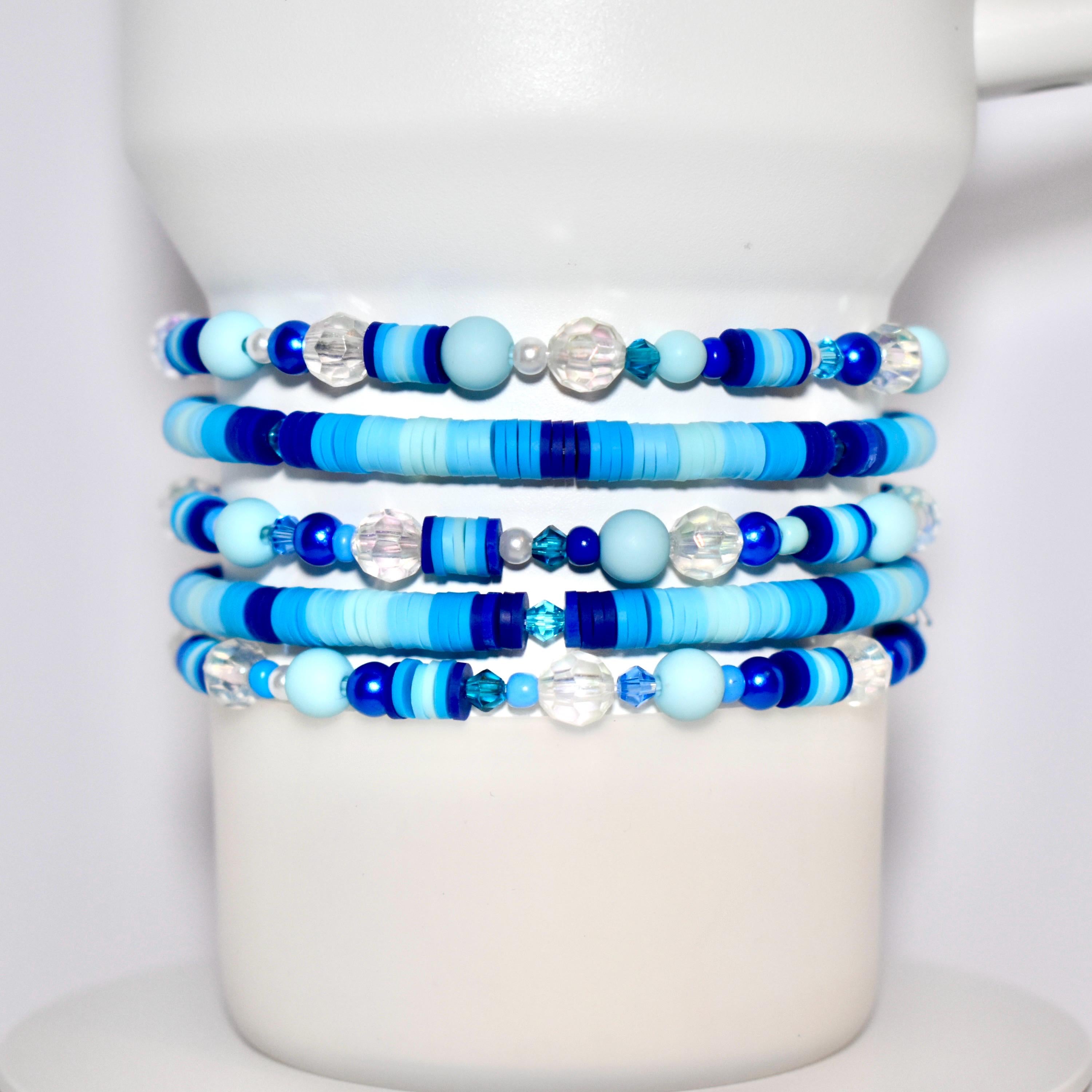 Stack of handmade beaded tumbler bracelets in various shades of blue, including matte, glossy, and translucent beads, wrapped around a white display stand. The Stanley bracelets feature round, disc, and faceted beads in a mix of light blue, dark blue, and clear tones.