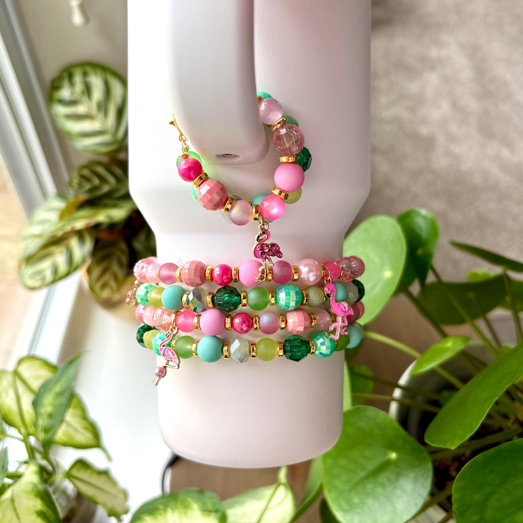 A set of five bracelets with pink, green, and gold beads. The bracelets are stacked on top of each other and have small flamingo charms hanging from them.