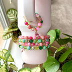 A set of five bracelets with pink, green, and gold beads. The bracelets are stacked on top of each other and have small flamingo charms hanging from them.