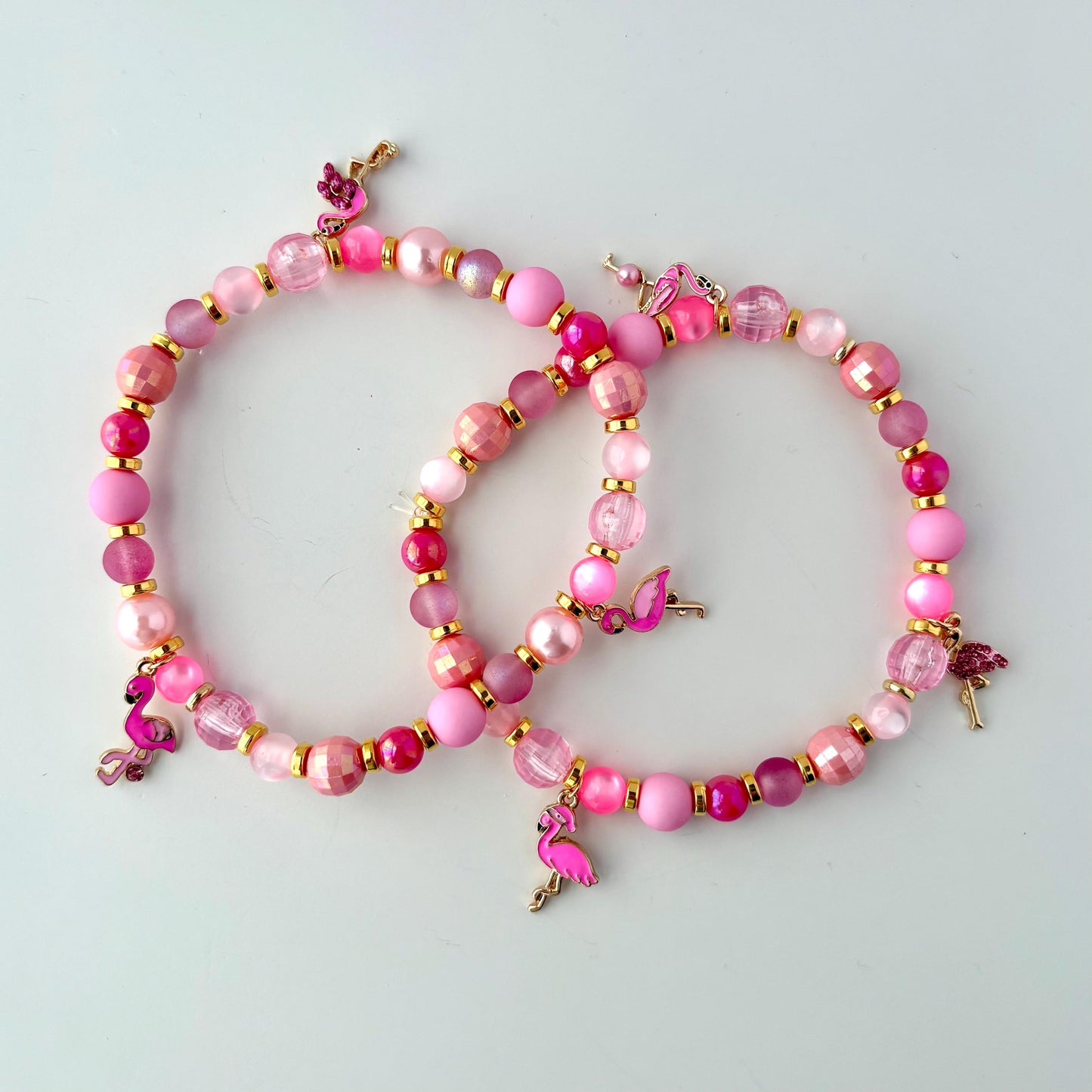 Two pink beaded bracelets with pink flamingo charms. The bracelets are made of pink and gold beads. Styled as vibrant tropical accessories for drink cups or tumblers.