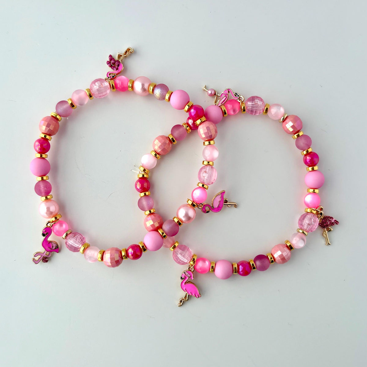 Two pink beaded bracelets with pink flamingo charms. The bracelets are made of pink and gold beads. Styled as vibrant tropical accessories for drink cups or tumblers.