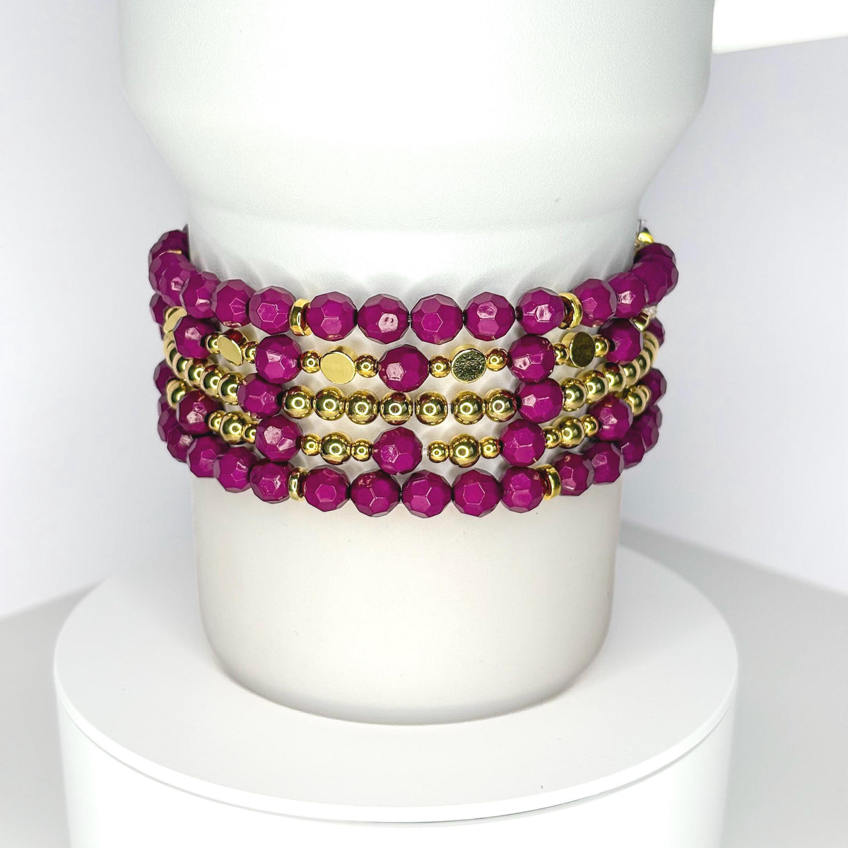 Handmade tumbler bracelet set featuring faceted magenta beads and shiny gold accents, stacked around a white tumbler for a stylish, elegant look.