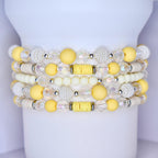 Stack of beaded bracelets in shades of yellow, white, and clear. The bracelets feature a variety of bead types, including round, textured, and iridescent beads. The bracelets are stacked on a white cylindrical surface.