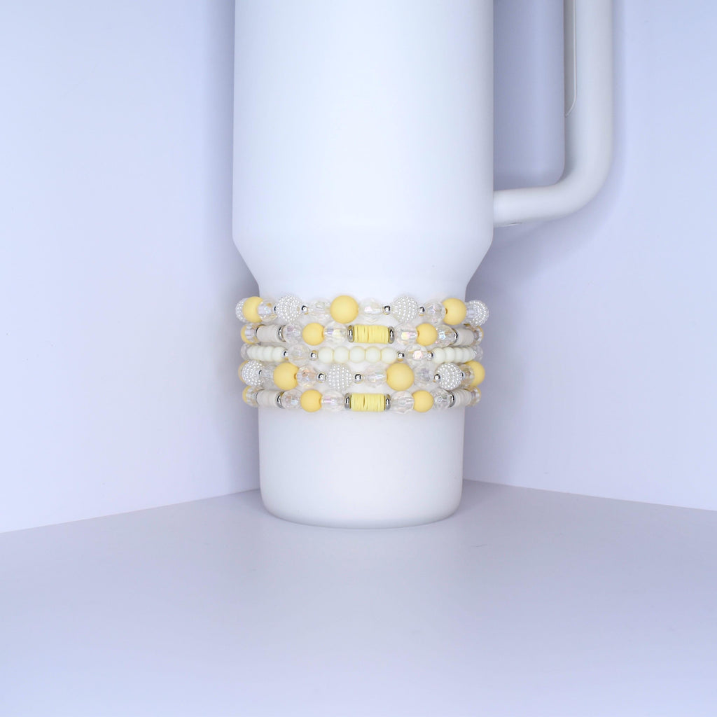 A white tumbler with a handle is adorned with several beaded bracelets. The bracelets feature a mix of yellow, white, and clear beads. Some beads are round, while others are faceted, creating a textured and colorful accessory.