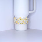 A white tumbler with a handle is adorned with several beaded bracelets. The bracelets feature a mix of yellow, white, and clear beads. Some beads are round, while others are faceted, creating a textured and colorful accessory.