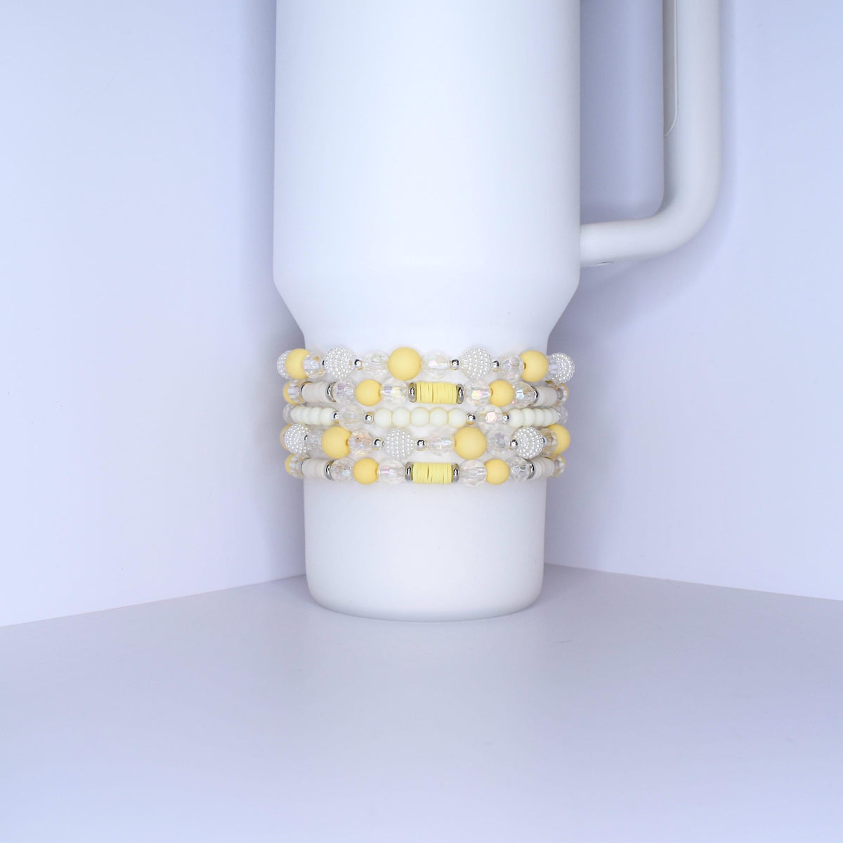 A white tumbler with a handle is adorned with several beaded bracelets. The bracelets feature a mix of yellow, white, and clear beads. Some beads are round, while others are faceted, creating a textured and colorful accessory.