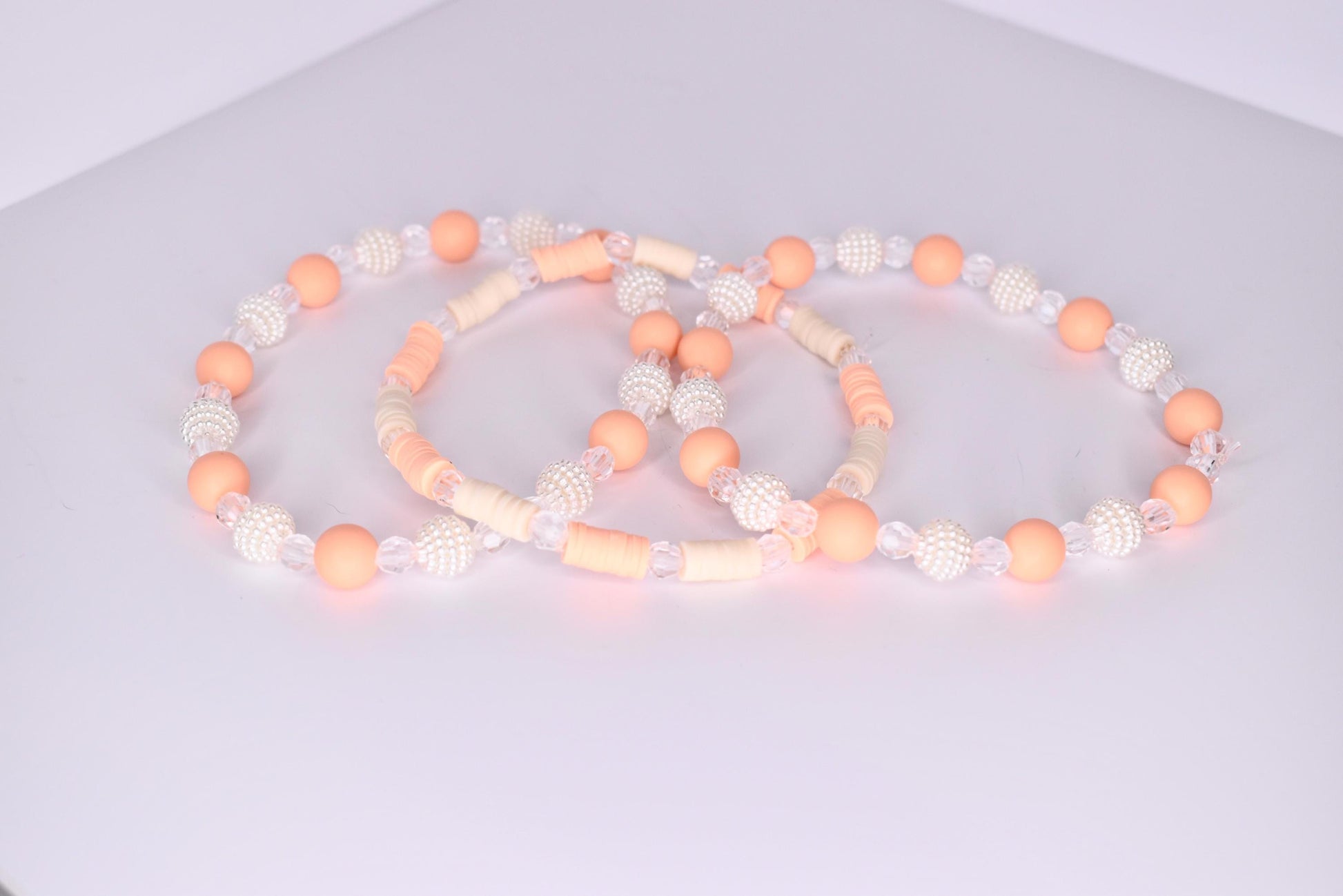 A trio of tumbler bracelets arranged in an overlapping display on a white surface, featuring a mix of peach matte beads, clear faceted beads, textured white pearl beads, and flat cream and peach disc beads in complementary patterns.