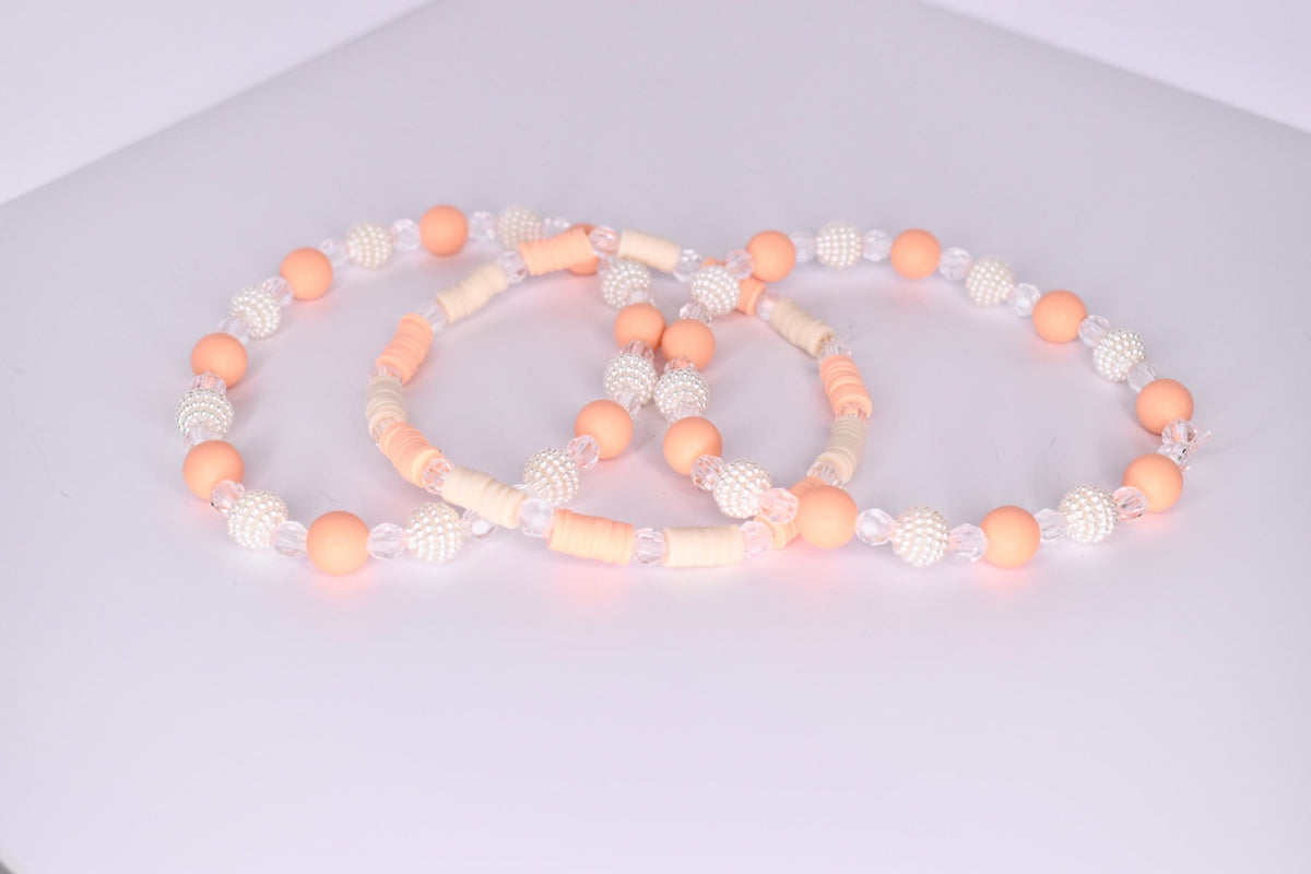 A trio of tumbler bracelets arranged in an overlapping display on a white surface, featuring a mix of peach matte beads, clear faceted beads, textured white pearl beads, and flat cream and peach disc beads in complementary patterns.