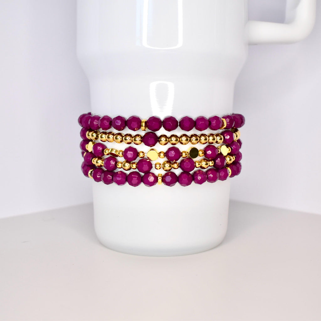 A set of five bracelets featuring a mix of deep purple and gold beads. The bracelets are stacked on top of each other, creating a layered look.