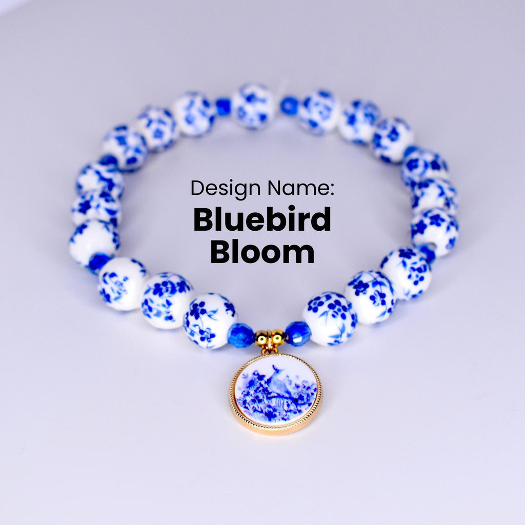 A white and blue ceramic necklace with a bluebird pendant. The necklace is made of round beads with a floral design. The pendant is a round disc with a bluebird and flowers in a blue and white design.