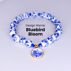 A white and blue ceramic necklace with a bluebird pendant. The necklace is made of round beads with a floral design. The pendant is a round disc with a bluebird and flowers in a blue and white design.