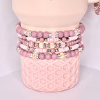 A collection of stacked bracelets in varying shades of pink. The bracelets feature a mix of round beads, sparkling rhinestone accents, and flat disc-shaped beads. The bracelets are displayed on a light pink, textured cylindrical base.
