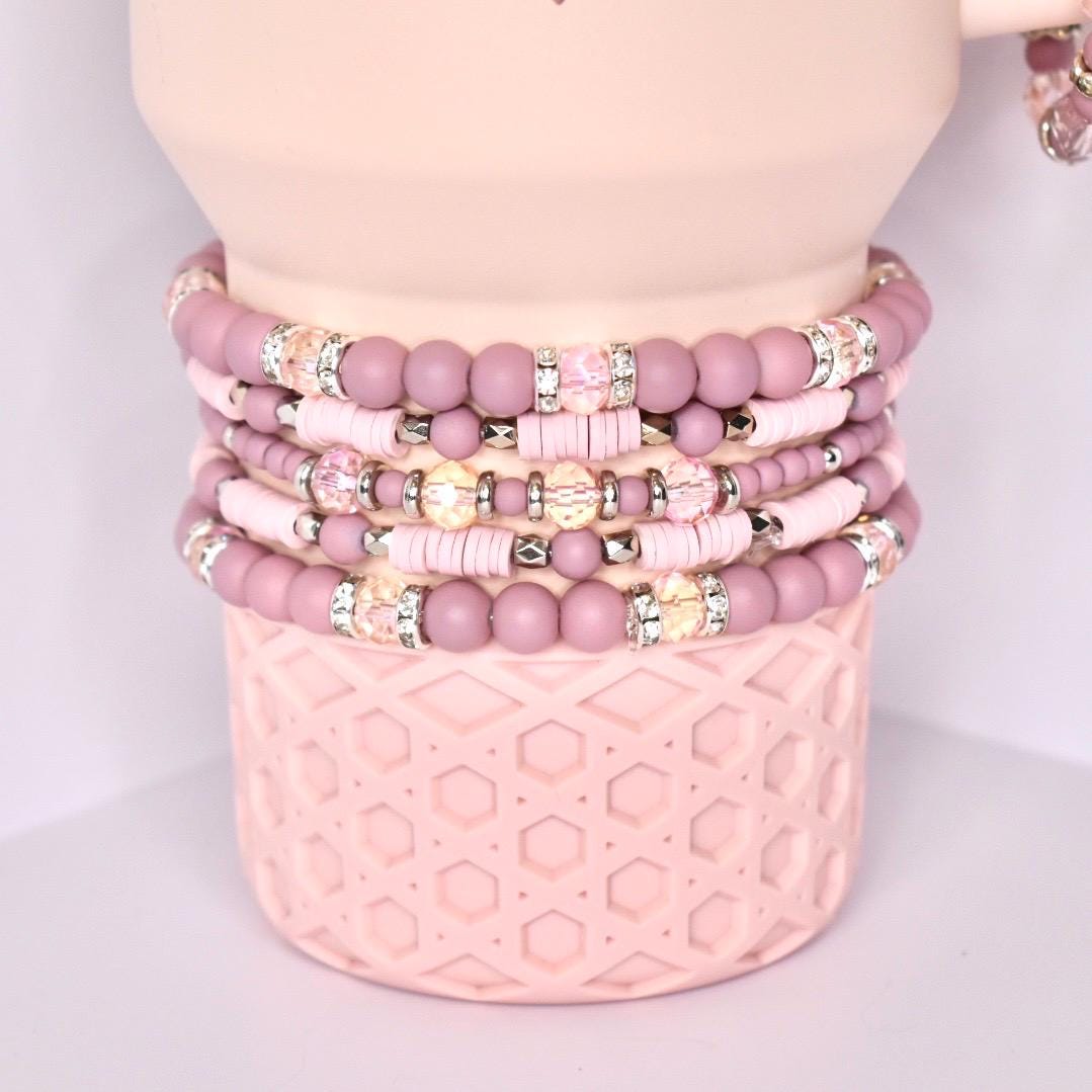 A collection of stacked bracelets in varying shades of pink. The bracelets feature a mix of round beads, sparkling rhinestone accents, and flat disc-shaped beads. The bracelets are displayed on a light pink, textured cylindrical base.