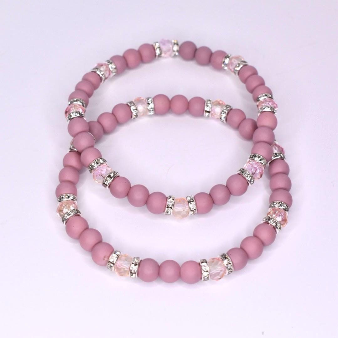 Two beaded bracelets in a dusty rose color. Each bracelet features small, round beads and sections of larger, clear beads with silver accents. The bracelets are stacked together, showcasing their similar design and color palette.