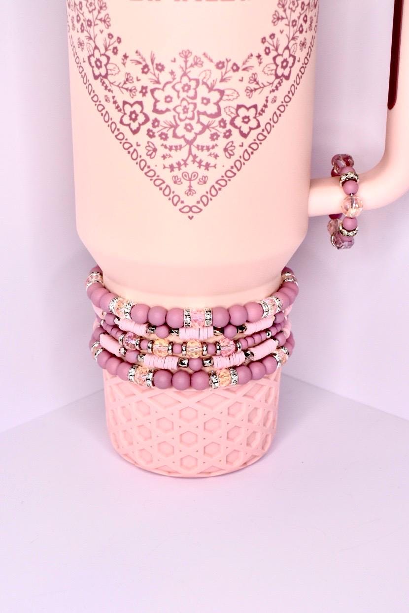 A stack of pink beaded bracelets and a pink tumbler. The bracelets are various shades of pink with silver accents. The tumbler is light pink with a floral design and a handle. A smaller bracelet is on the handle. The bracelets are stacked on a pink base.