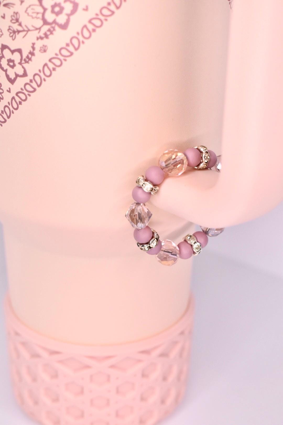 A beaded tumbler handle bracelet with a pink and clear color scheme. The ring features small, round pink beads interspersed with larger, faceted clear beads. Silver-colored, decorative accents are placed between some of the beads. The ring is displayed on a light pink surface.