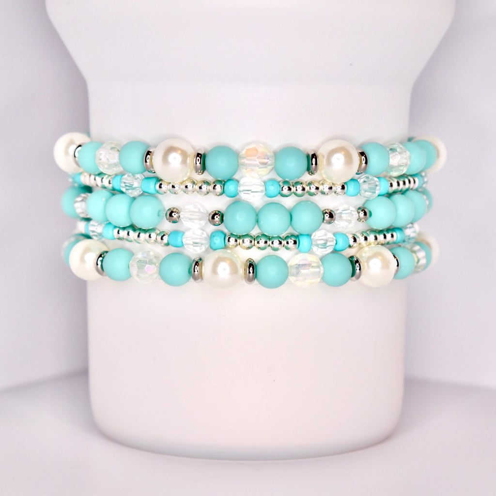 A close-up of a white tumbler with a handle, wrapped with five stacked beaded bracelets in a coordinated turquoise and white theme. The bracelets feature matte turquoise beads, iridescent clear beads, pearl-like white beads, and shiny silver spacers.