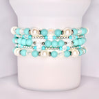 A close-up of a white tumbler with a handle, wrapped with five stacked beaded bracelets in a coordinated turquoise and white theme. The bracelets feature matte turquoise beads, iridescent clear beads, pearl-like white beads, and shiny silver spacers.