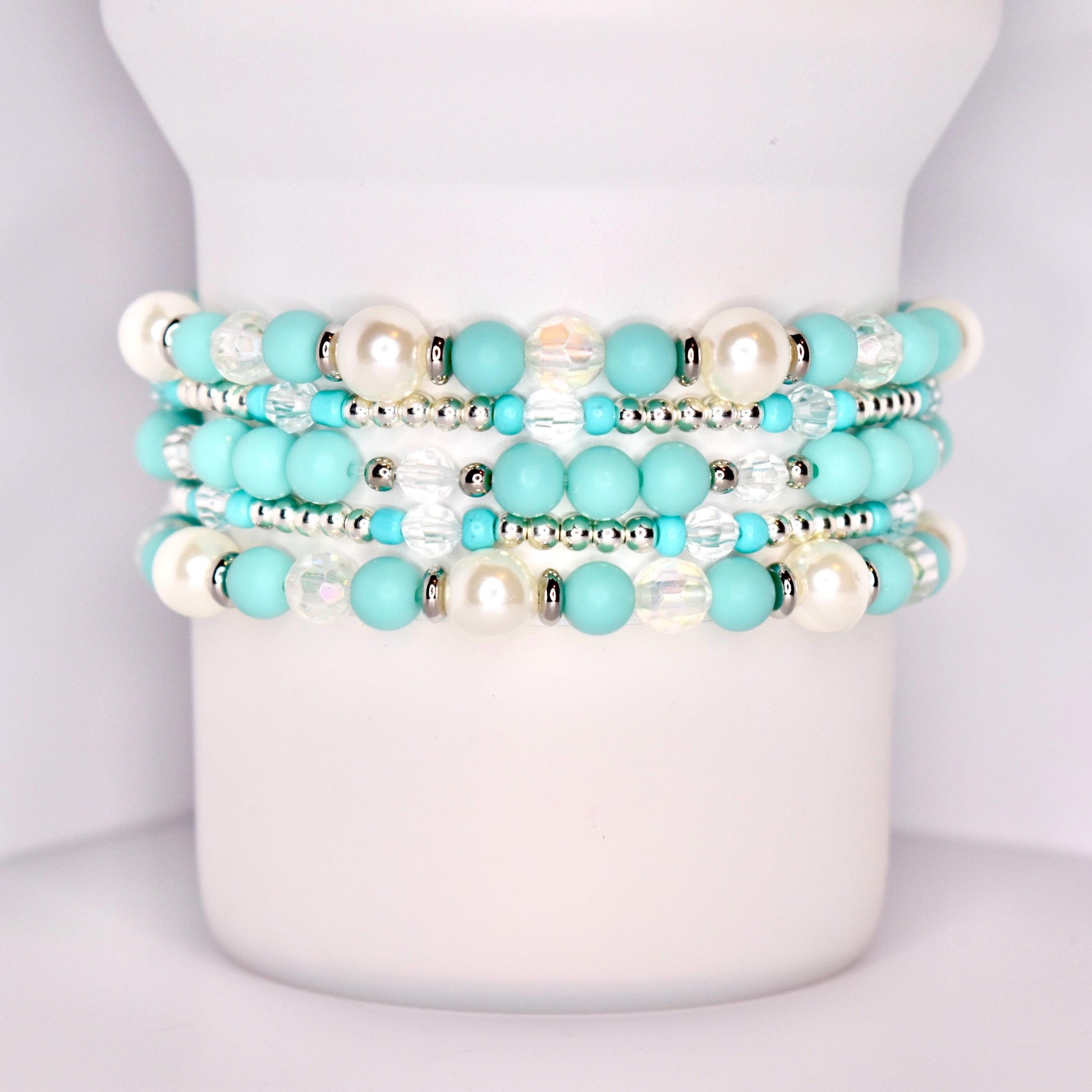 A close-up of a white tumbler with a handle, wrapped with five stacked beaded bracelets in a coordinated turquoise and white theme. The bracelets feature matte turquoise beads, iridescent clear beads, pearl-like white beads, and shiny silver spacers.