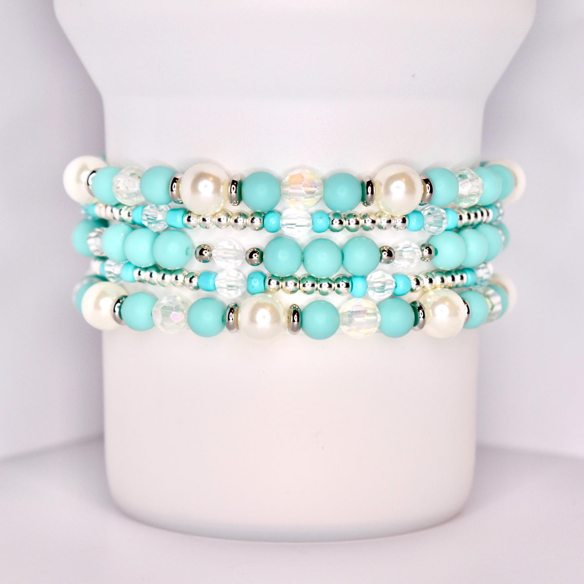 A close-up of a white tumbler with a handle, wrapped with five stacked beaded bracelets in a coordinated turquoise and white theme. The bracelets feature matte turquoise beads, iridescent clear beads, pearl-like white beads, and shiny silver spacers.