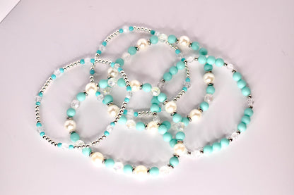 Five beaded tumbler bracelets with a turquoise and white color scheme arranged loosely on a light surface. The bracelets include a mix of matte turquoise beads, pearl-like white beads, clear faceted beads, and silver spacers, showing their individual designs.
