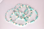 Five beaded tumbler bracelets with a turquoise and white color scheme arranged loosely on a light surface. The bracelets include a mix of matte turquoise beads, pearl-like white beads, clear faceted beads, and silver spacers, showing their individual designs.