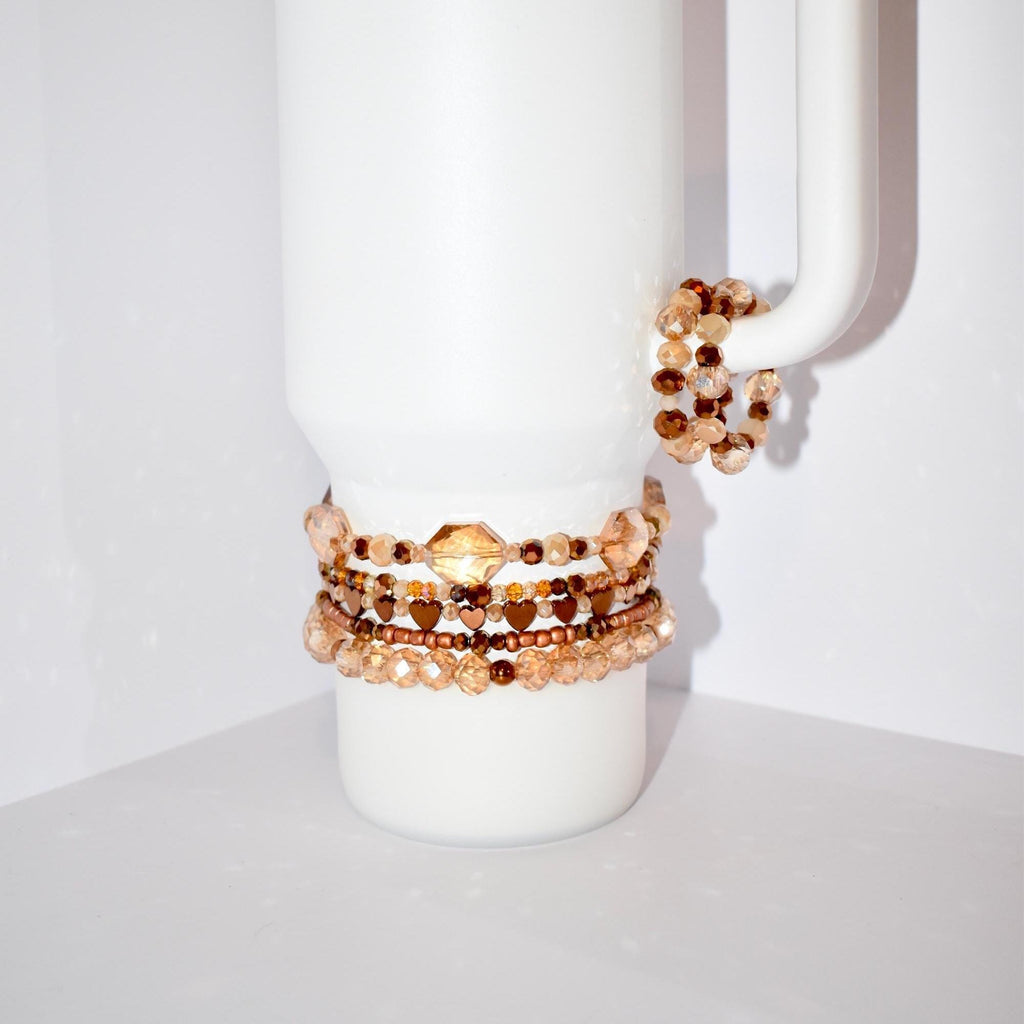 Several beaded bracelets in shades of brown and gold are displayed on a white ceramic mug. The bracelets feature various bead sizes and shapes, including heart-shaped beads. One bracelet is wrapped around the mug's handle.