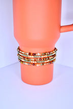 A pumpkin spice tumbler with a handle is adorned with multiple beaded bracelets. The bracelets are wrapped around the tumbler, showcasing a variety of small, colorful beads in shades of gold, brown, green, and red. The bracelets appear to be handmade.