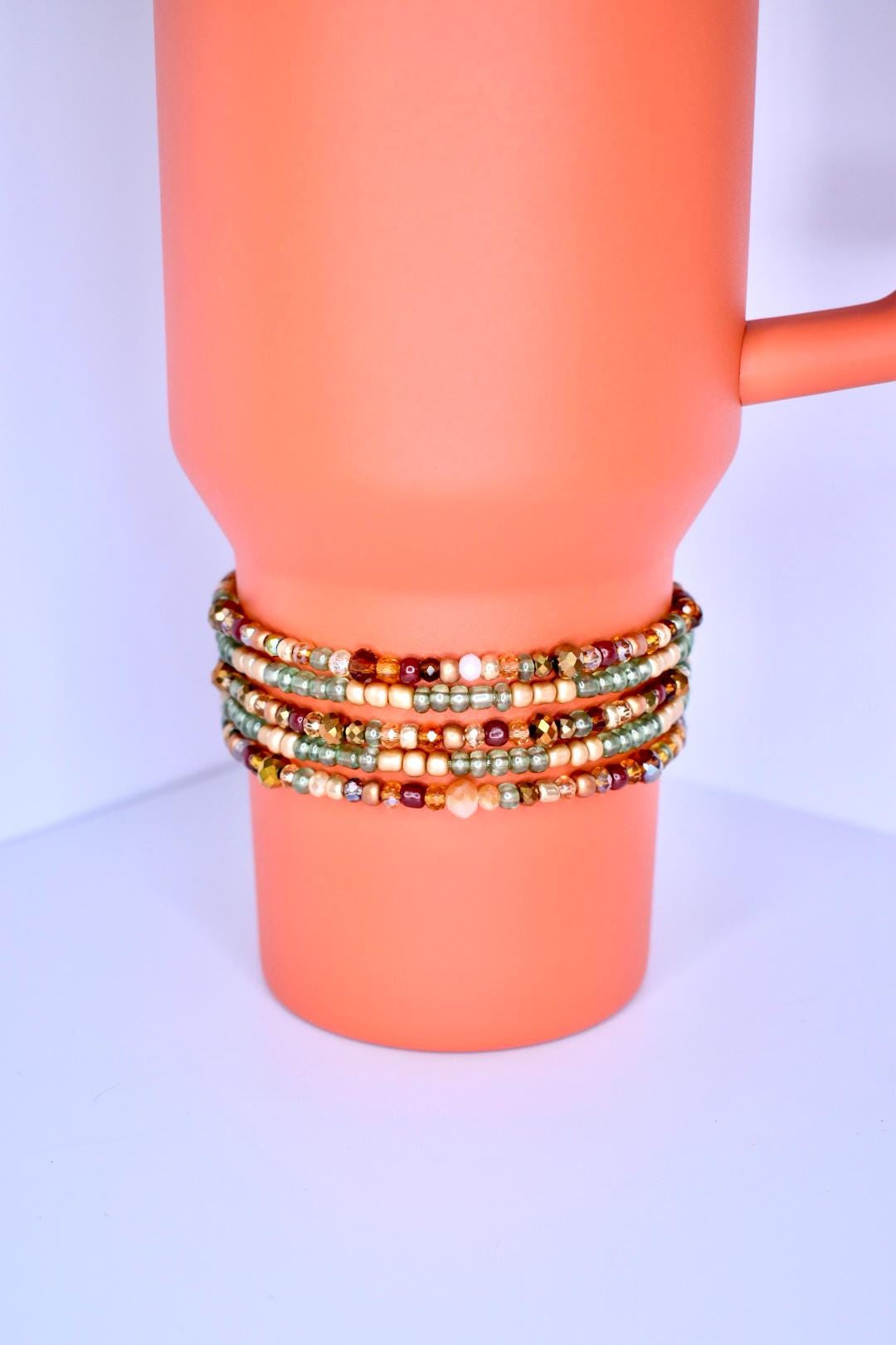 A pumpkin spice tumbler with a handle is adorned with multiple beaded bracelets. The bracelets are wrapped around the tumbler, showcasing a variety of small, colorful beads in shades of gold, brown, green, and red. The bracelets appear to be handmade.