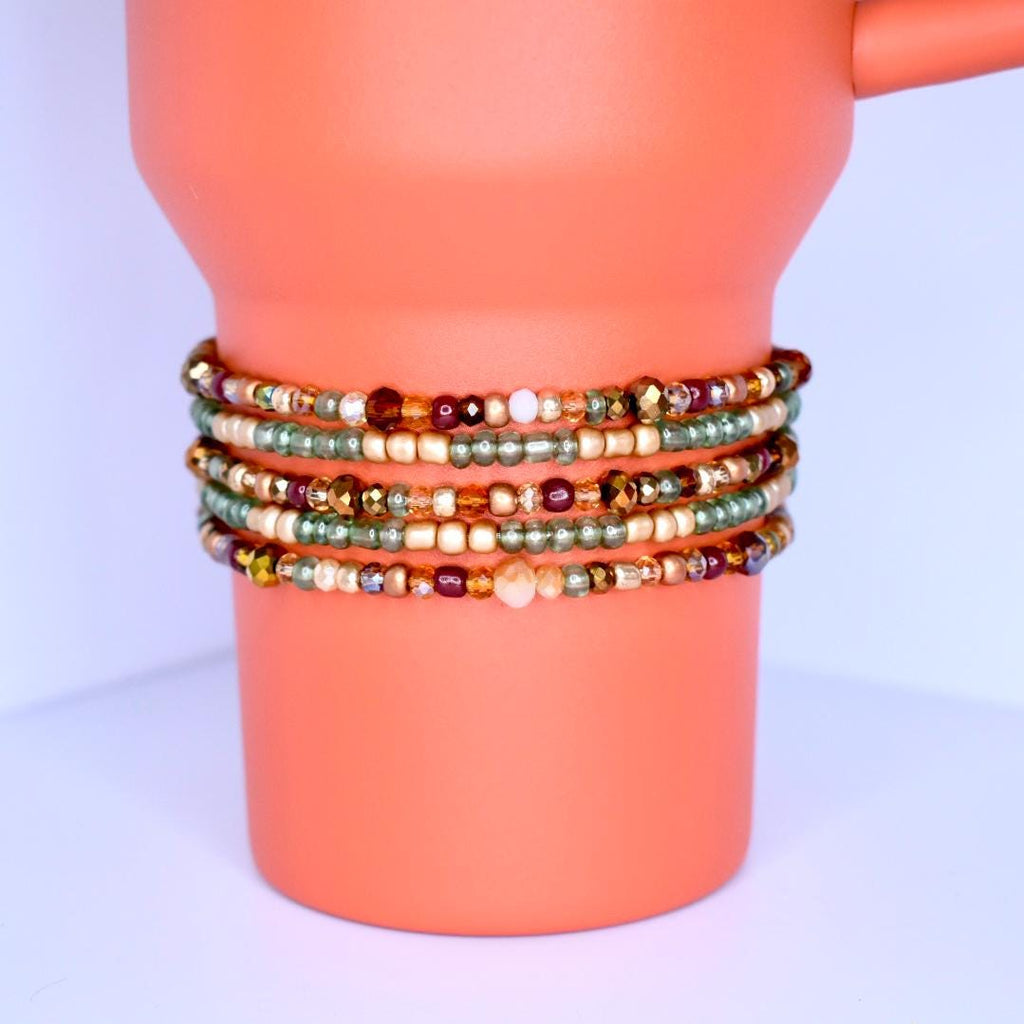 A stack of beaded bracelets in various colors. The bracelets are wrapped around a pumpkin spice Stanley. The beads are small and come in shades of gold, green, brown, and cream. The bracelets appear to be handmade and have a delicate, intricate design.