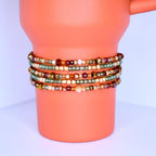 A stack of beaded bracelets in various colors. The bracelets are wrapped around a pumpkin spice Stanley. The beads are small and come in shades of gold, green, brown, and cream. The bracelets appear to be handmade and have a delicate, intricate design.