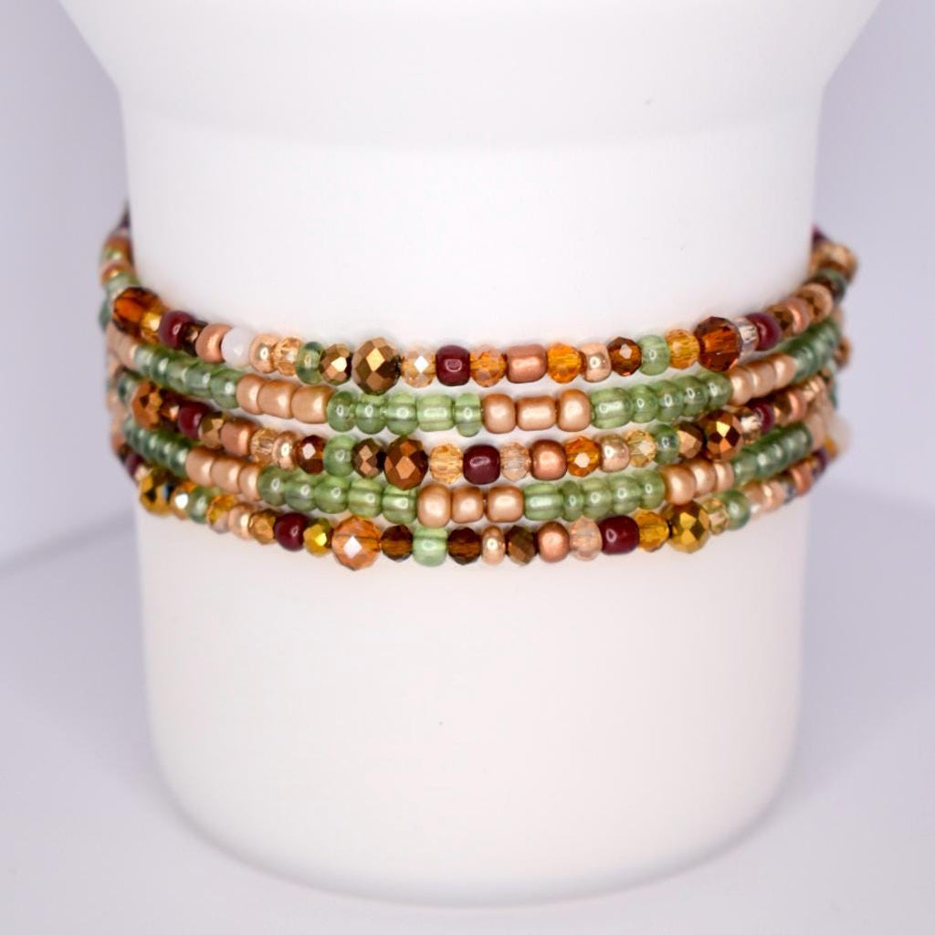 A stack of beaded bracelets in shades of green, brown, and gold. The bracelets are wrapped around a white cylindrical object. The beads vary in size and shape, with some faceted and others round. The colors include olive green, amber, and copper, creating a warm, earthy palette.