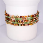 A stack of beaded bracelets in shades of green, brown, and gold. The bracelets are wrapped around a white cylindrical object. The beads vary in size and shape, with some faceted and others round. The colors include olive green, amber, and copper, creating a warm, earthy palette.
