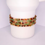 A collection of beaded tumbler bracelets in shades of green, gold, and brown. The bracelets are wrapped around a white cylindrical object. The beads vary in size and shape, creating a textured and colorful accessory.