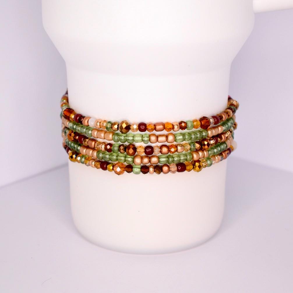 A collection of beaded tumbler bracelets in shades of green, gold, and brown. The bracelets are wrapped around a white cylindrical object. The beads vary in size and shape, creating a textured and colorful accessory.