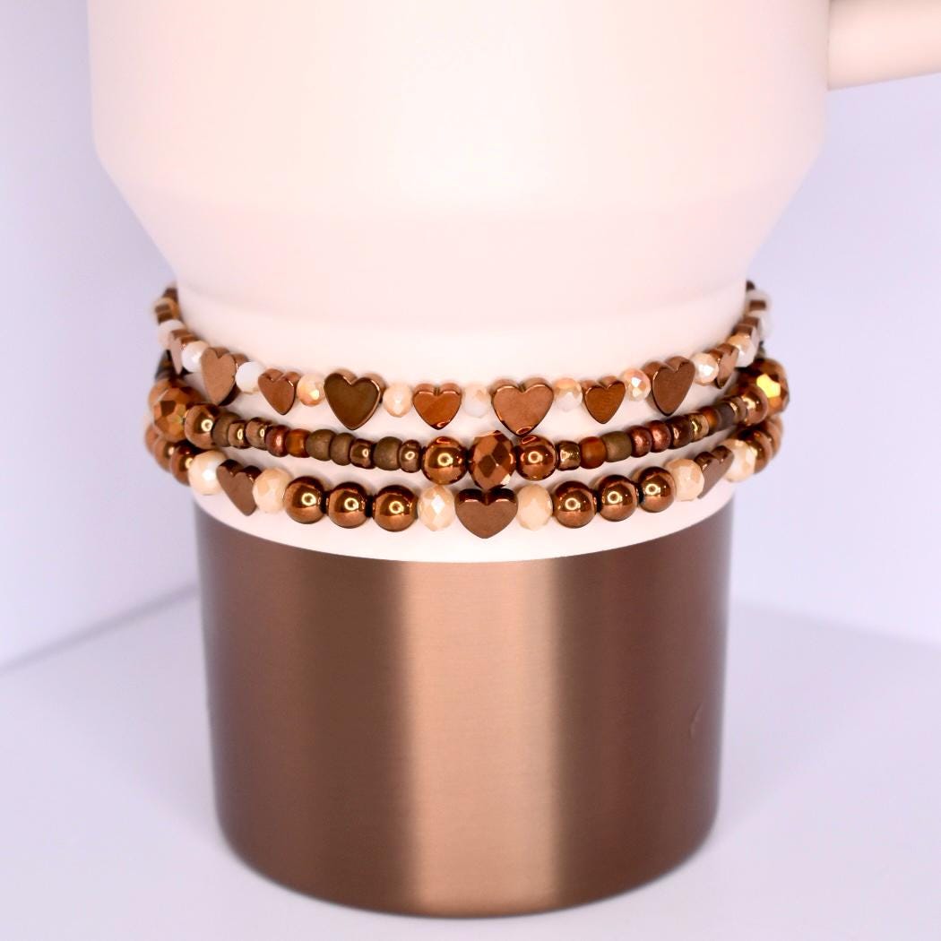 Three beaded bracelets in shades of brown and gold are stacked on a Gilded Sugar Stanley tumbler. The top bracelet features heart-shaped beads. The middle bracelet has small, round beads. The bottom bracelet has a mix of round and heart-shaped beads.