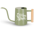 A sage green watering can with a long, curved spout and a wooden handle. The watering can features the words 'let love bloom' in white script with a floral design. The handle is a light brown color, and the can has a cylindrical body.