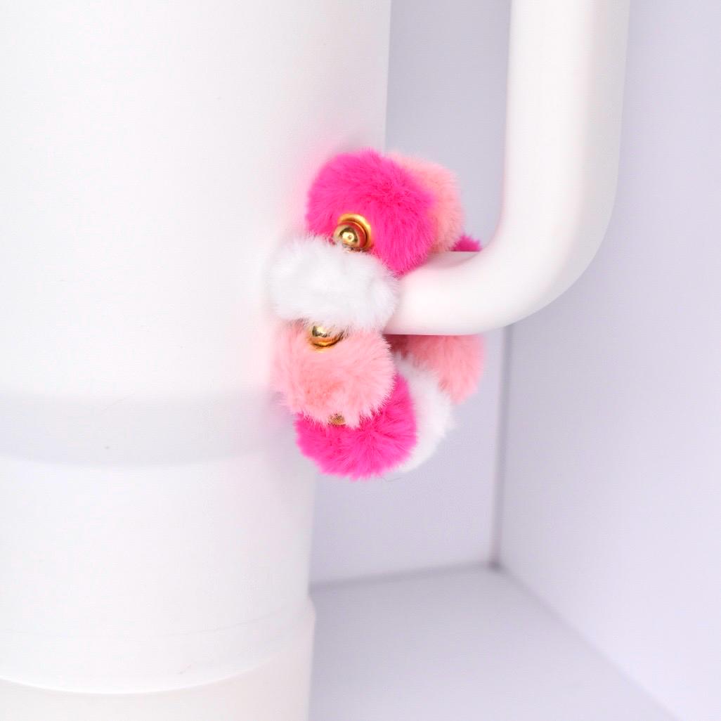 A colorful scrunchie with a mix of pink, white, and peach-colored faux fur pom-poms. The scrunchie is accented with small gold-colored metal details. The accessory is designed to wrap around a tumbler handle.